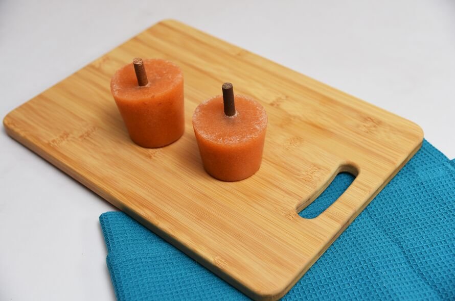 Orange-colourd dog-friendly ice lolly with a dog chew for a stick standing on a chopping board